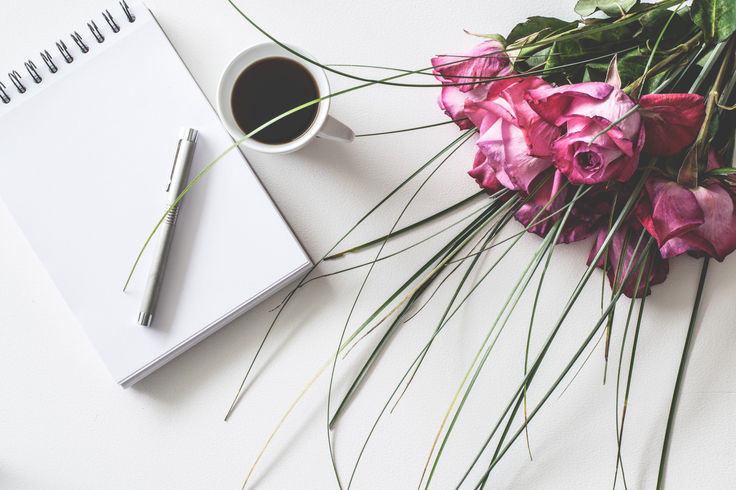 Notepad, pen, mug, and flower bouquet on white surface.