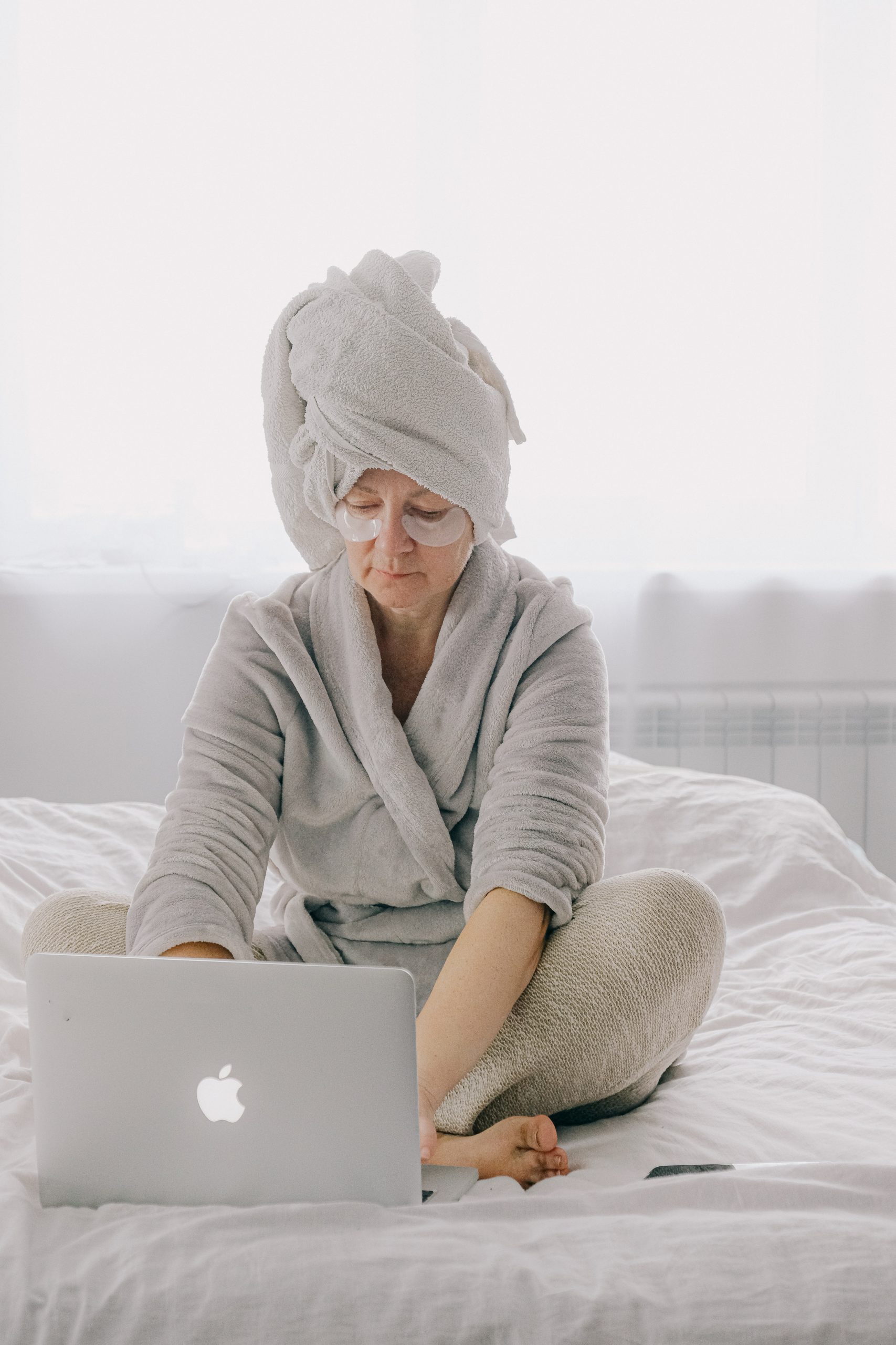Woman sitting on bed with eyepatches on while on laptop.