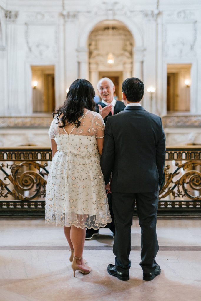 Bride and groom holding hands in front of officiant in building.
