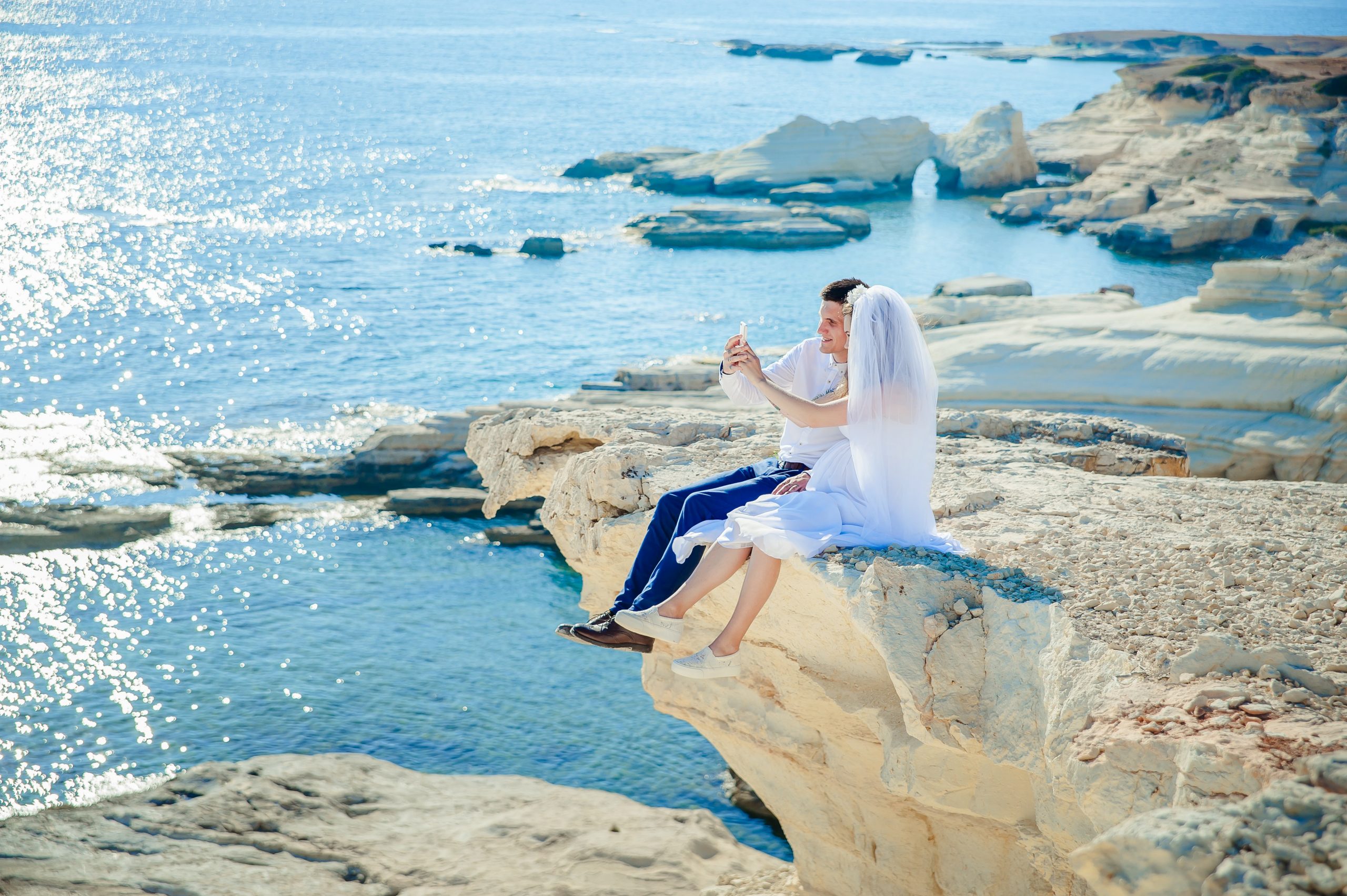 couple in weddingwear sitting on cliff above tropical water