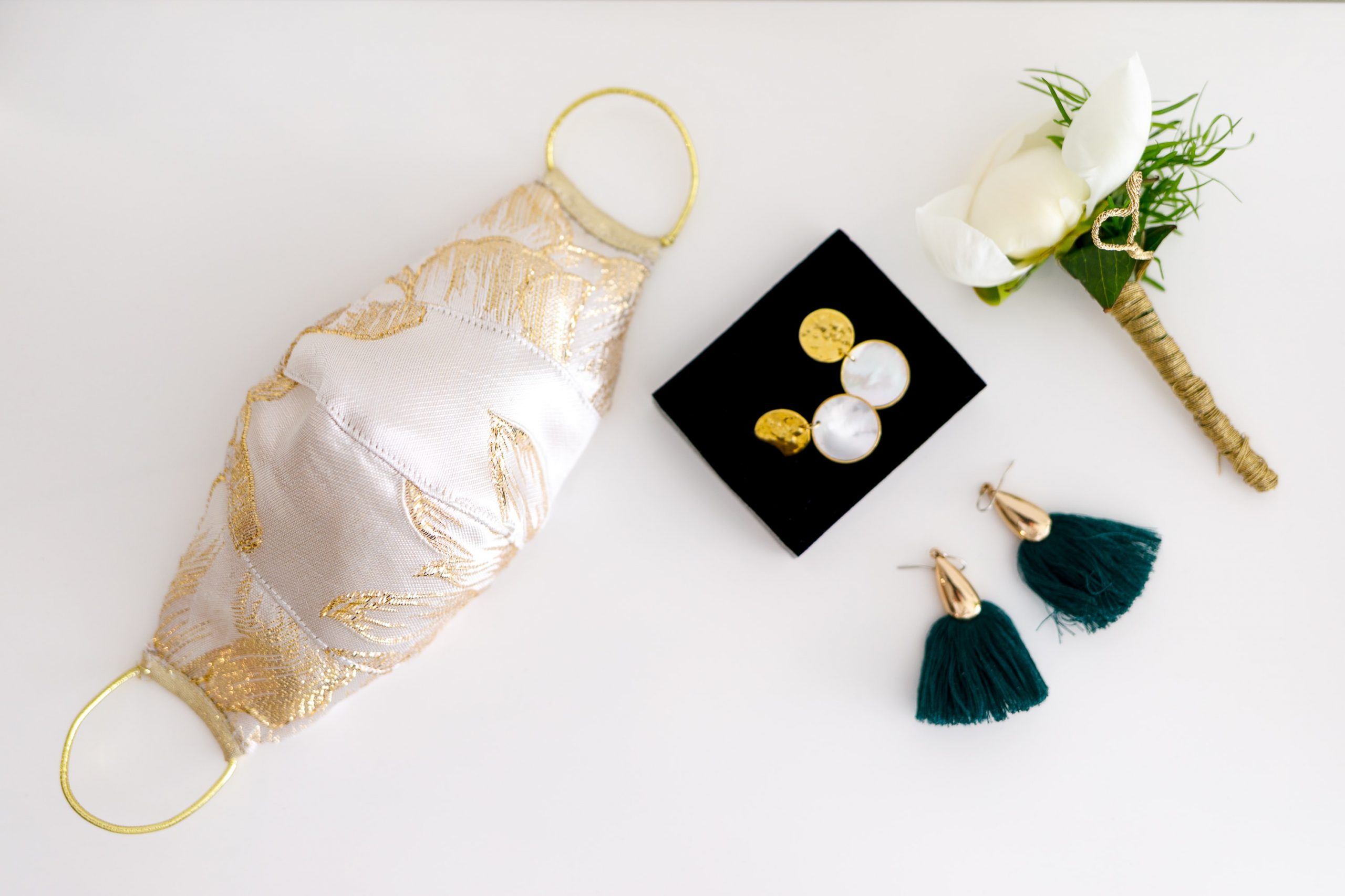 A bridal photo with an elegantly embroided white facemask layed out next to earrings and a boutonniere